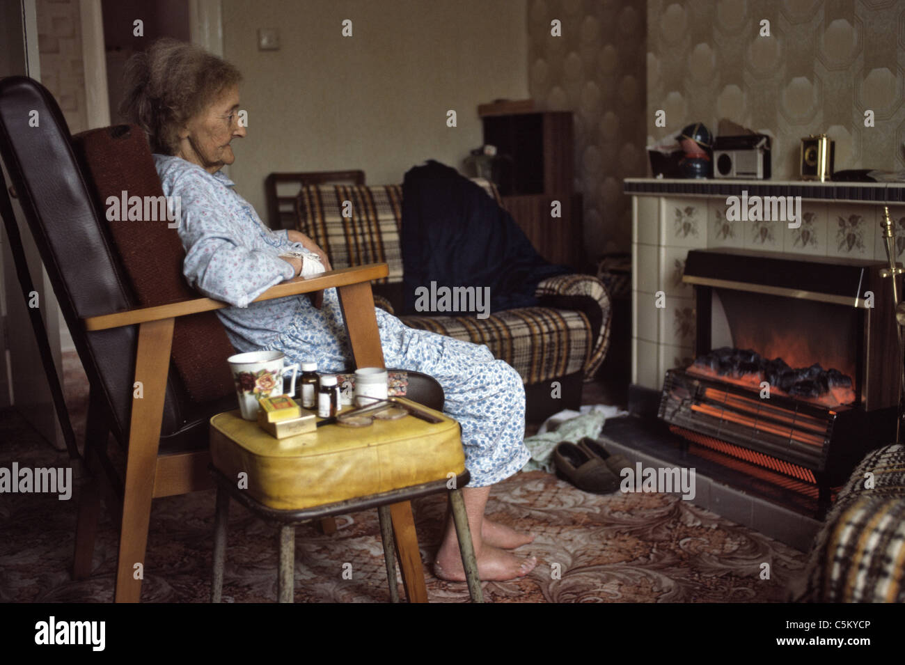 Molto vecchia donna fragile seduti davanti al fuoco elettrico Foto Stock
