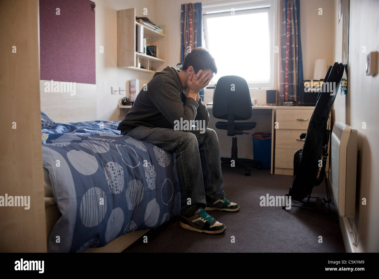Giovane studente caucasico maschio. Testa nelle mani, seduto su un letto, solo in camera lontano da casa - homesick. Foto Stock