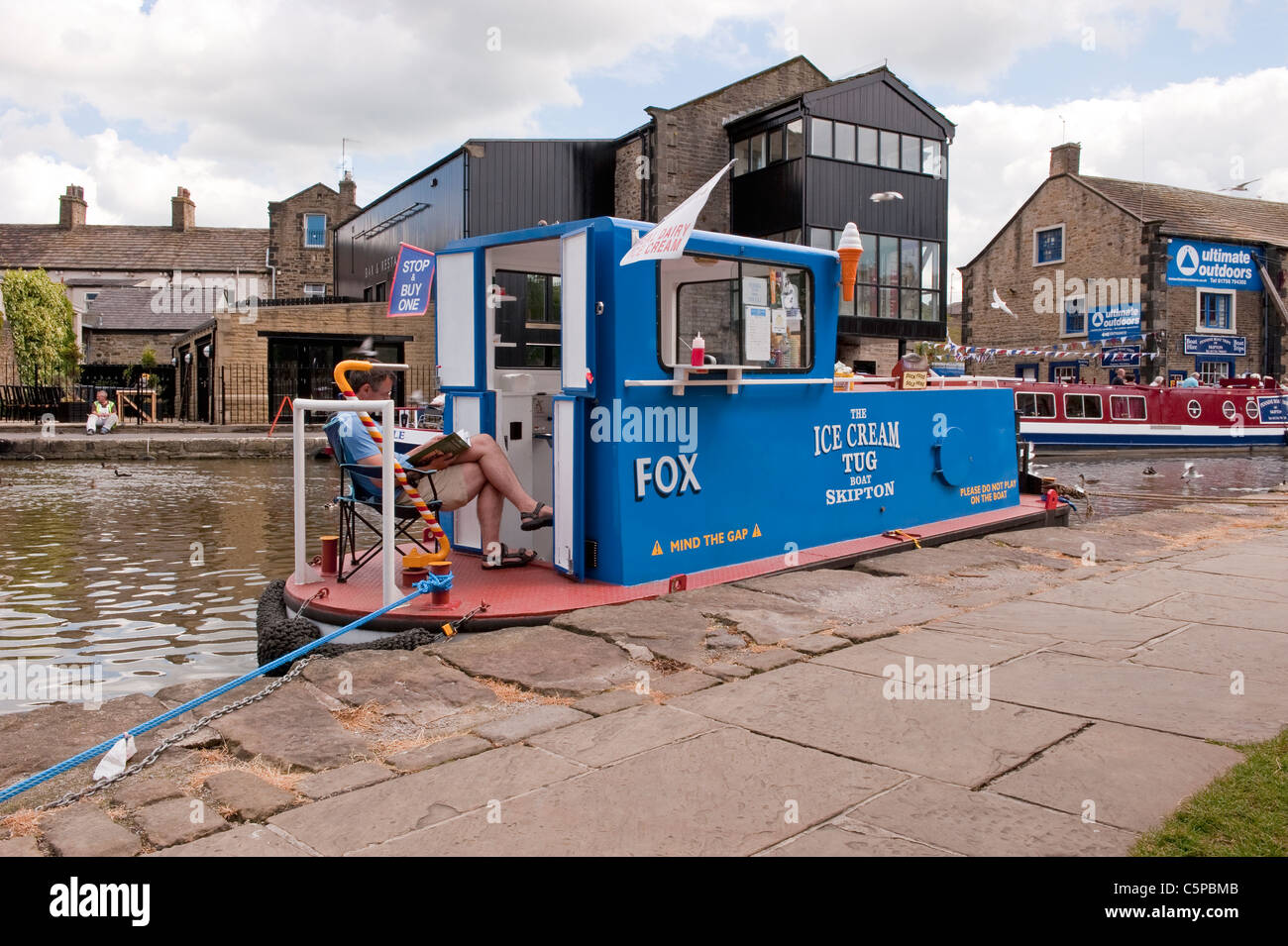 Gelateria 'Fox' (rimorchiatore convertito) che vende gelati ormeggiati da un alzaia e uomo seduto sul ponte di lettura - Leeds Liverpool Canal, Skipton, Inghilterra, UK. Foto Stock