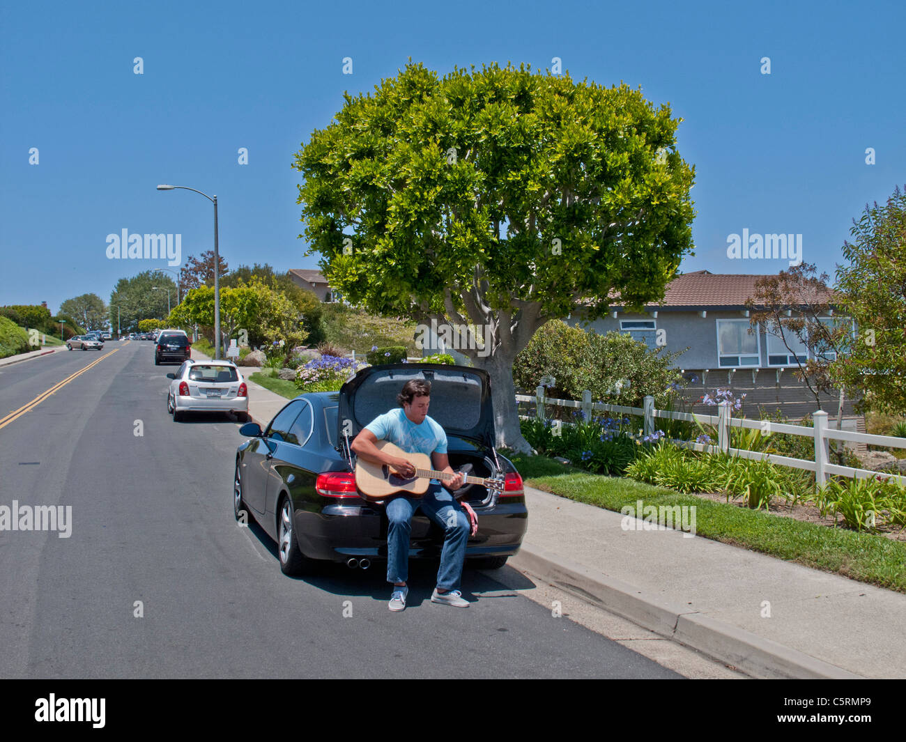 Seduto sulla parte posteriore della sua vettura sotto un sole cielo blu, un giovane uomo di pratica con la sua chitarra su un soleggiato suburban street in Laguna Nig Foto Stock