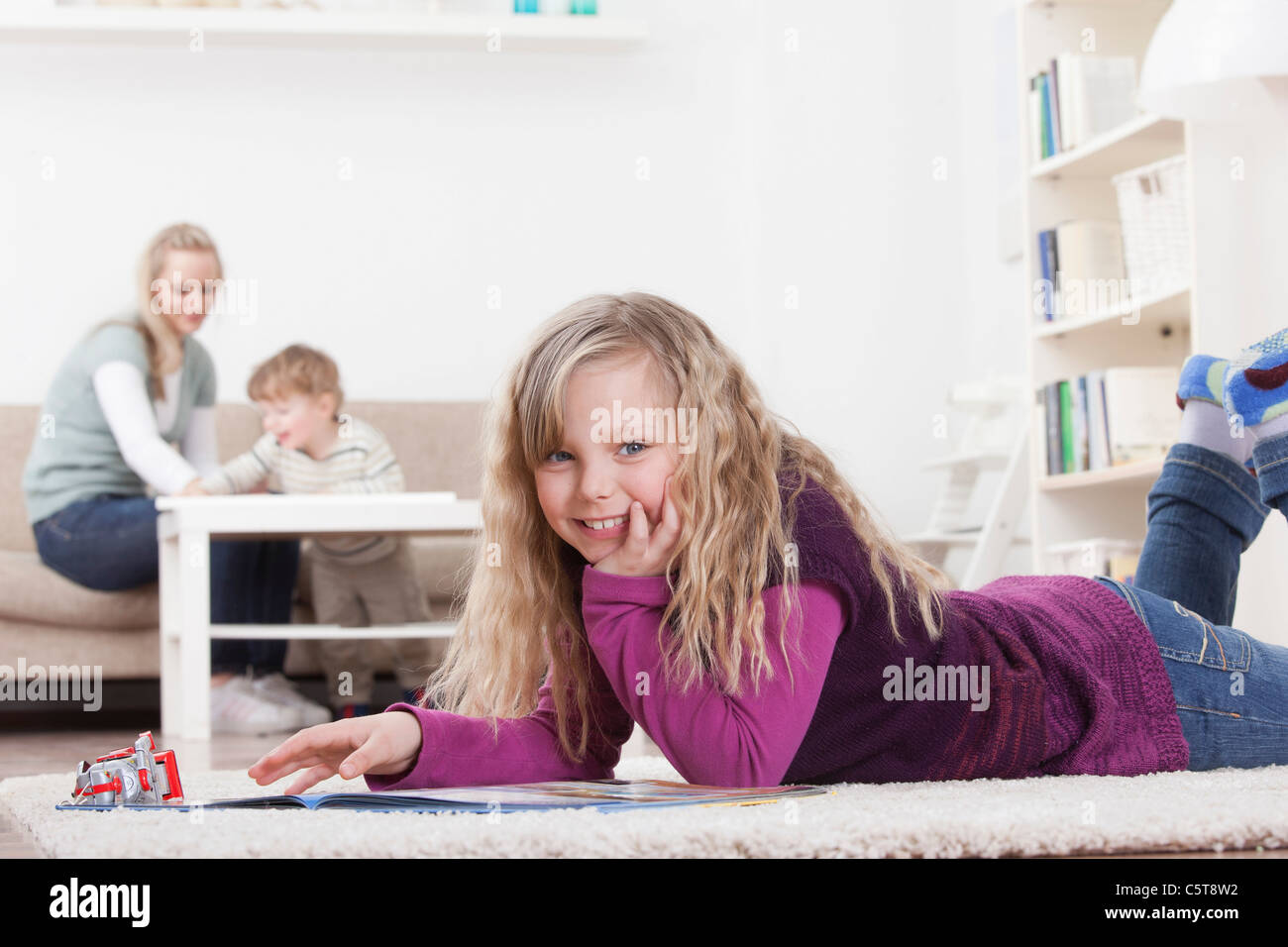 In Germania, in Baviera, Monaco di Baviera, ragazza con libro sul pavimento, madre e figlio in background Foto Stock