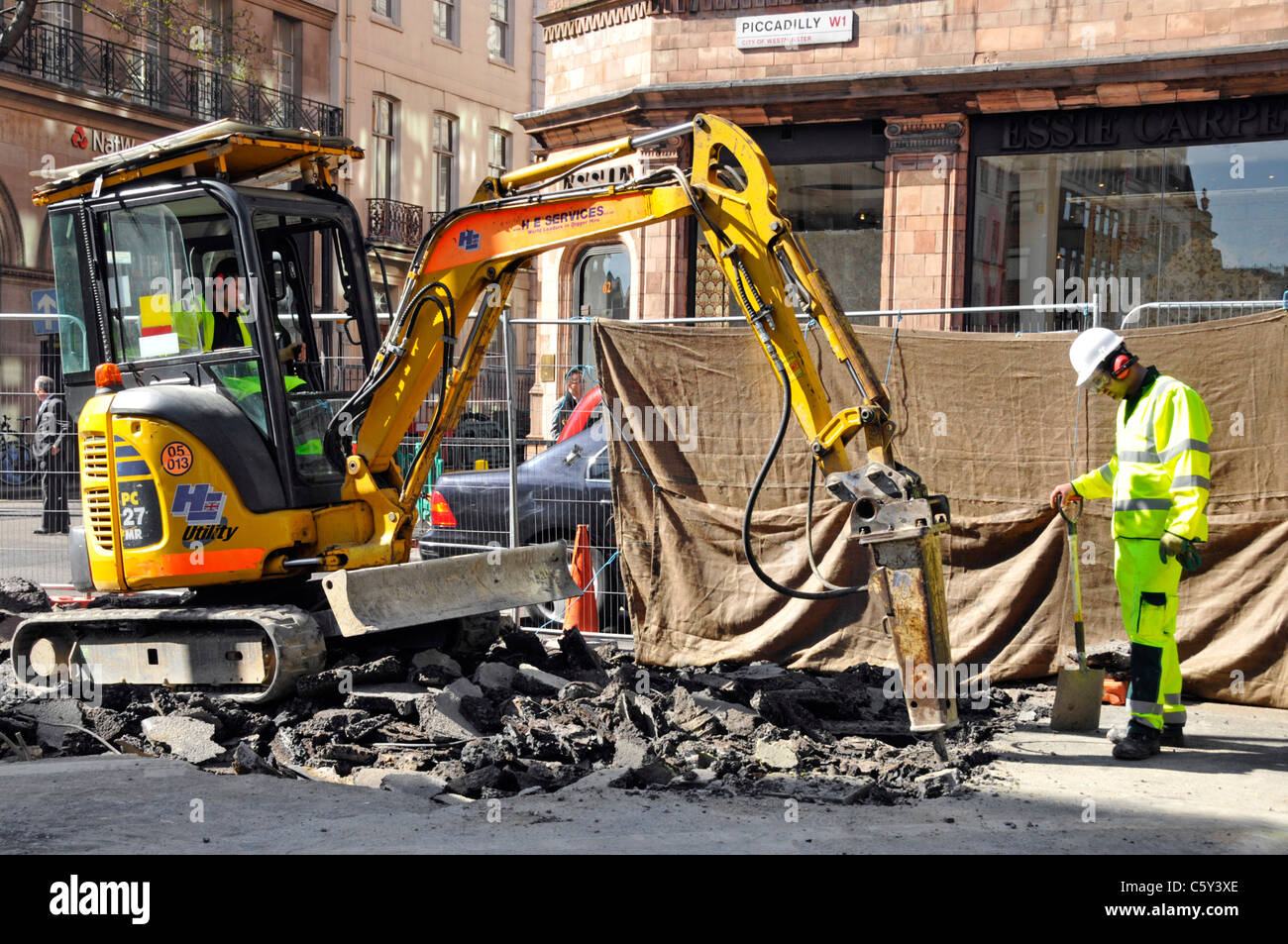 Macchina escavatore con retro mini-scavatore dotata di demolitore pneumatico rumoroso per i palchi stradali a Piccadilly Londra Inghilterra UK Foto Stock