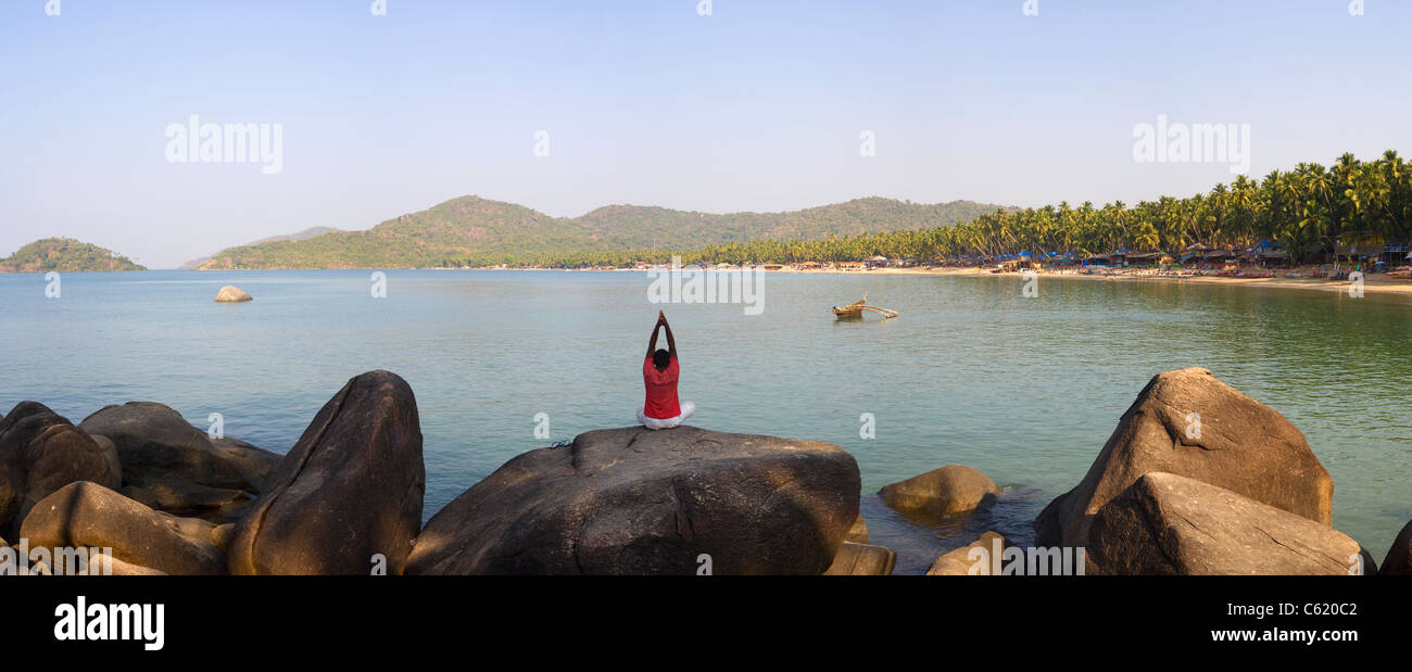 Un uomo indiano esegue lo yoga su una roccia presso la popolare località turistica della destinazione di Palolem spiaggia sul Mare Arabico, Goa, India Foto Stock