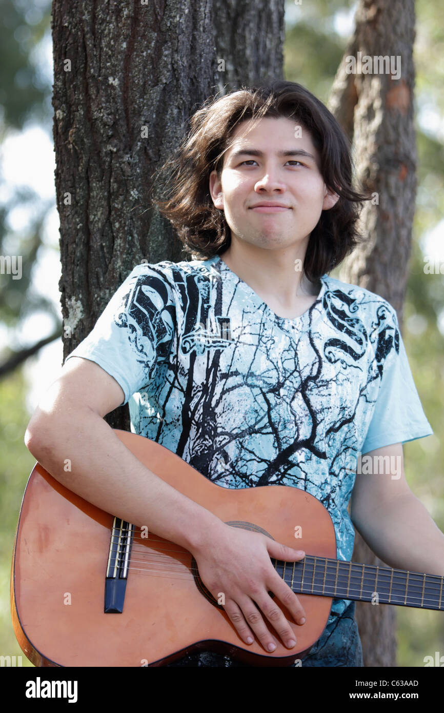 Un sorriso misto messicano ragazzo con i capelli lunghi e di suonare una chitarra con alberi in background Foto Stock