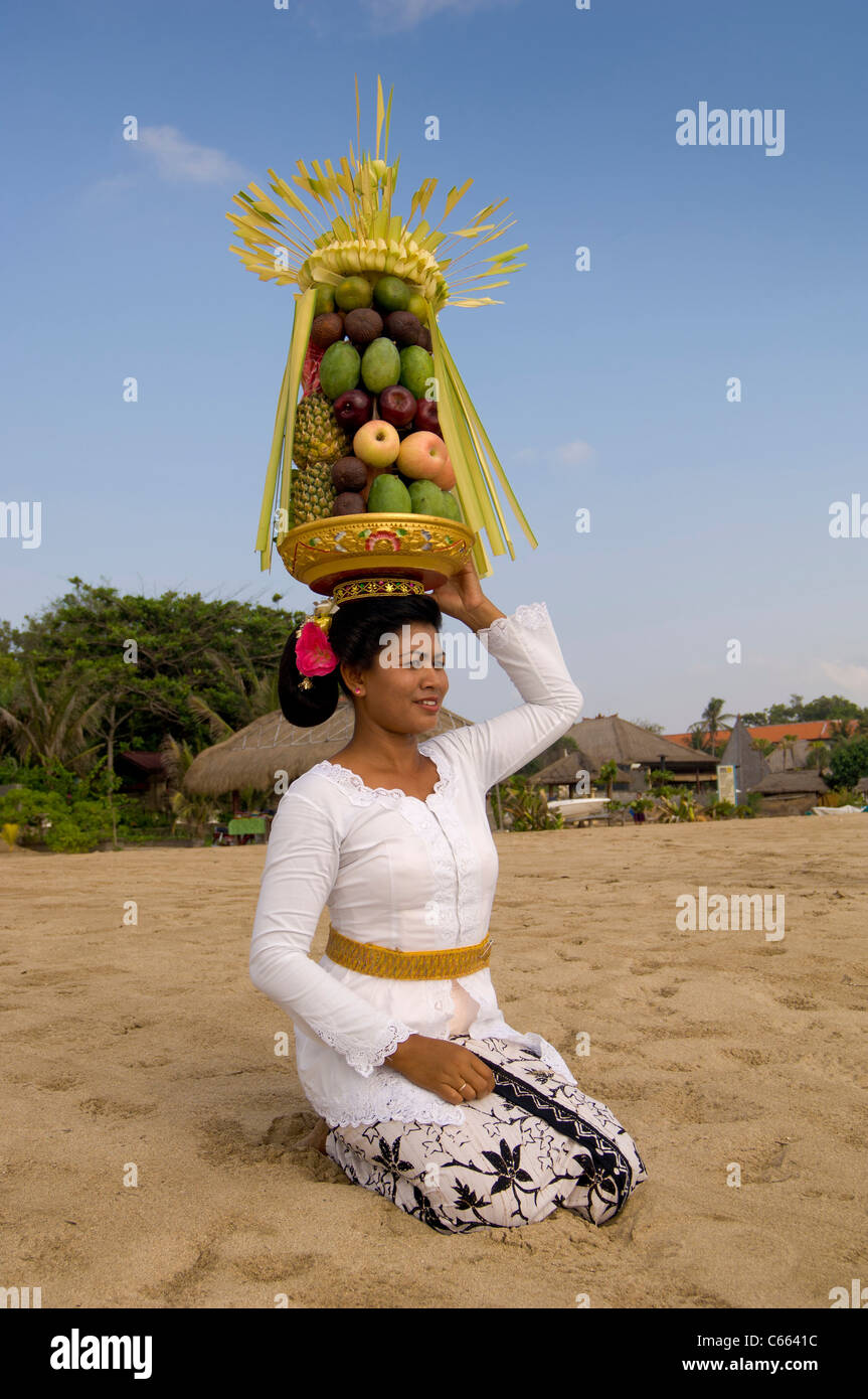 Donna balinese in costume tradizionale inginocchiata sulla spiaggia che porta un cestino di frutta, conosciuta anche come canang sari sulla sua testa. Foto Stock