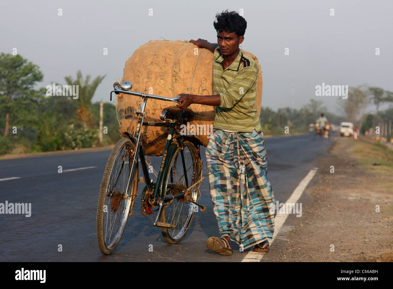 Giovane uomo lotte con sacco pesante in bicicletta fino una trafficata autostrada al tramonto nel Bengala Occidentale in India Foto Stock