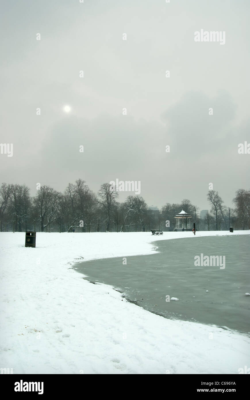 Pietra rotonda stagno in Hyde Park, Londra, ricoperto di ghiaccio con neve sul terreno attorno ad esso con la band stand in background Foto Stock