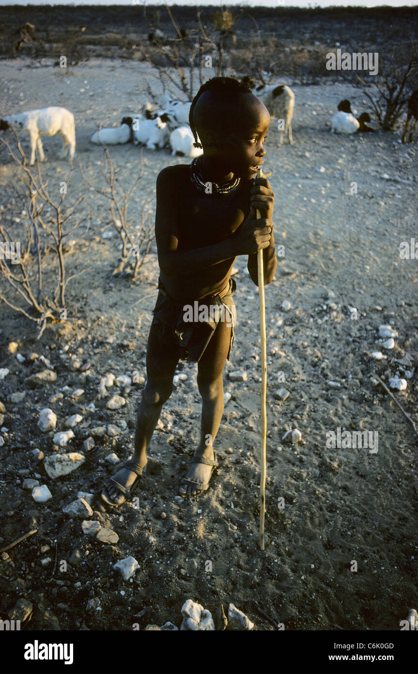 Giovane ragazzo Himba appoggiato su un bastone con capre in background Foto Stock