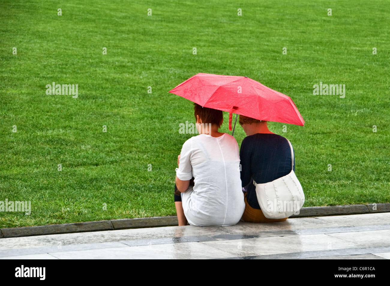 Due donne rifugiandosi sotto un ombrello rosso dalla pioggia. Vista dalla parte posteriore dietro. Close up Foto Stock