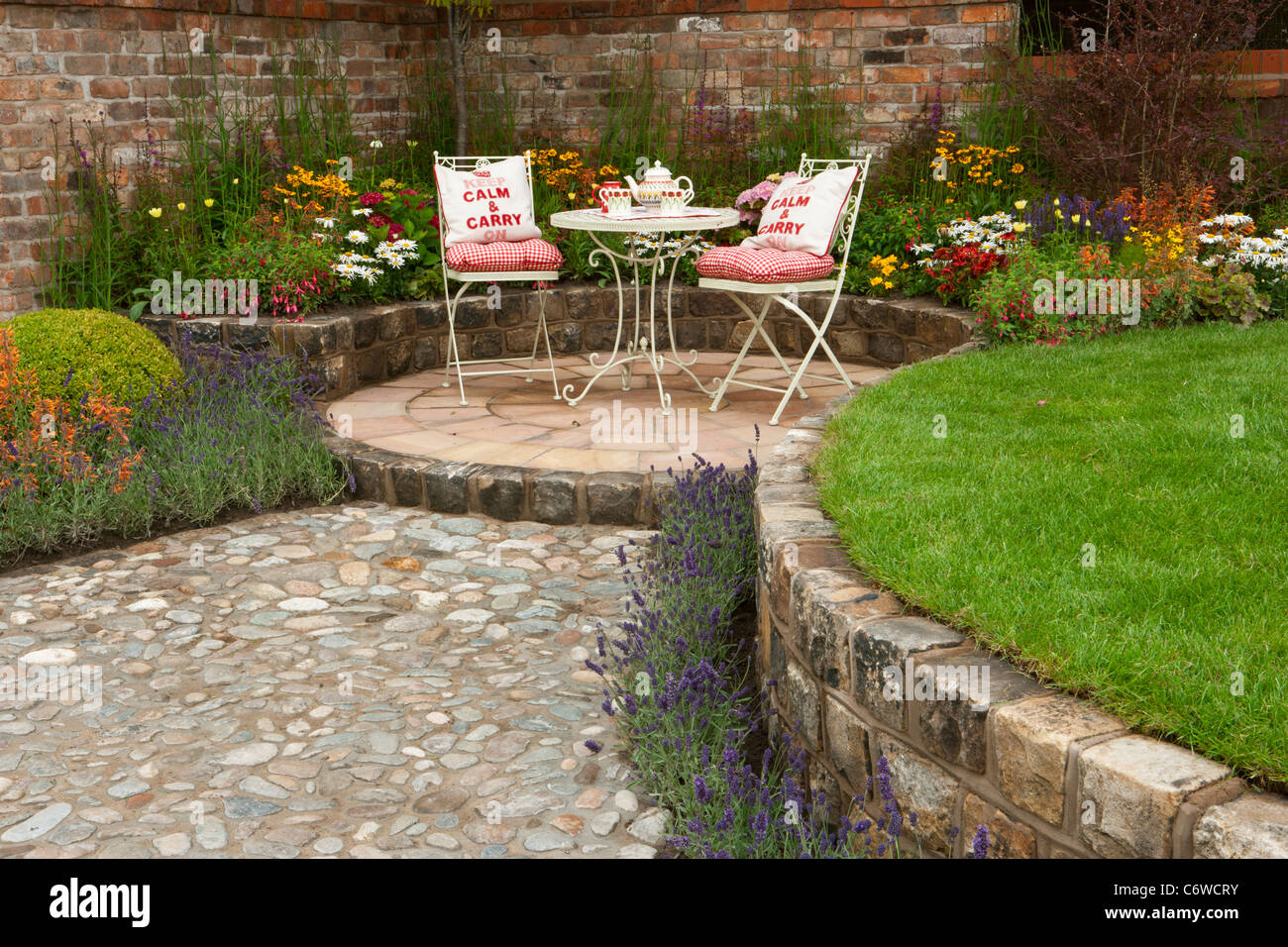 Piccolo patio circolare lastricato in pietra e prato con tavolo e sedie con posti a sedere con ciottoli sentiero di ciottoli e letti rialzati in mattoni rigenerati Foto Stock