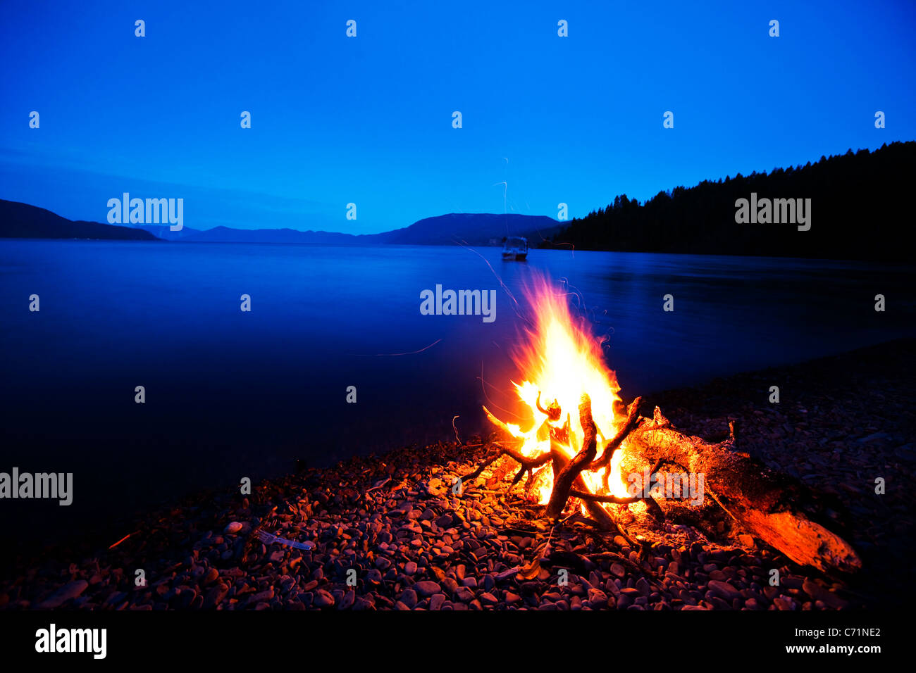 Un bellissimo campo di fuoco brucia al crepuscolo contro un profondo lago blu con una barca per wakeboard in background in Idaho. Foto Stock