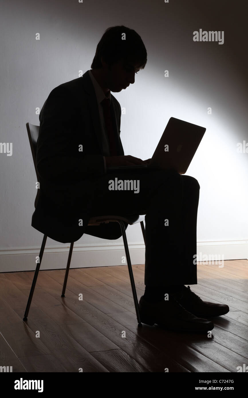 Silhouette di un giovane maschio indossa una tuta, seduta utilizzando un computer portatile. Foto Stock