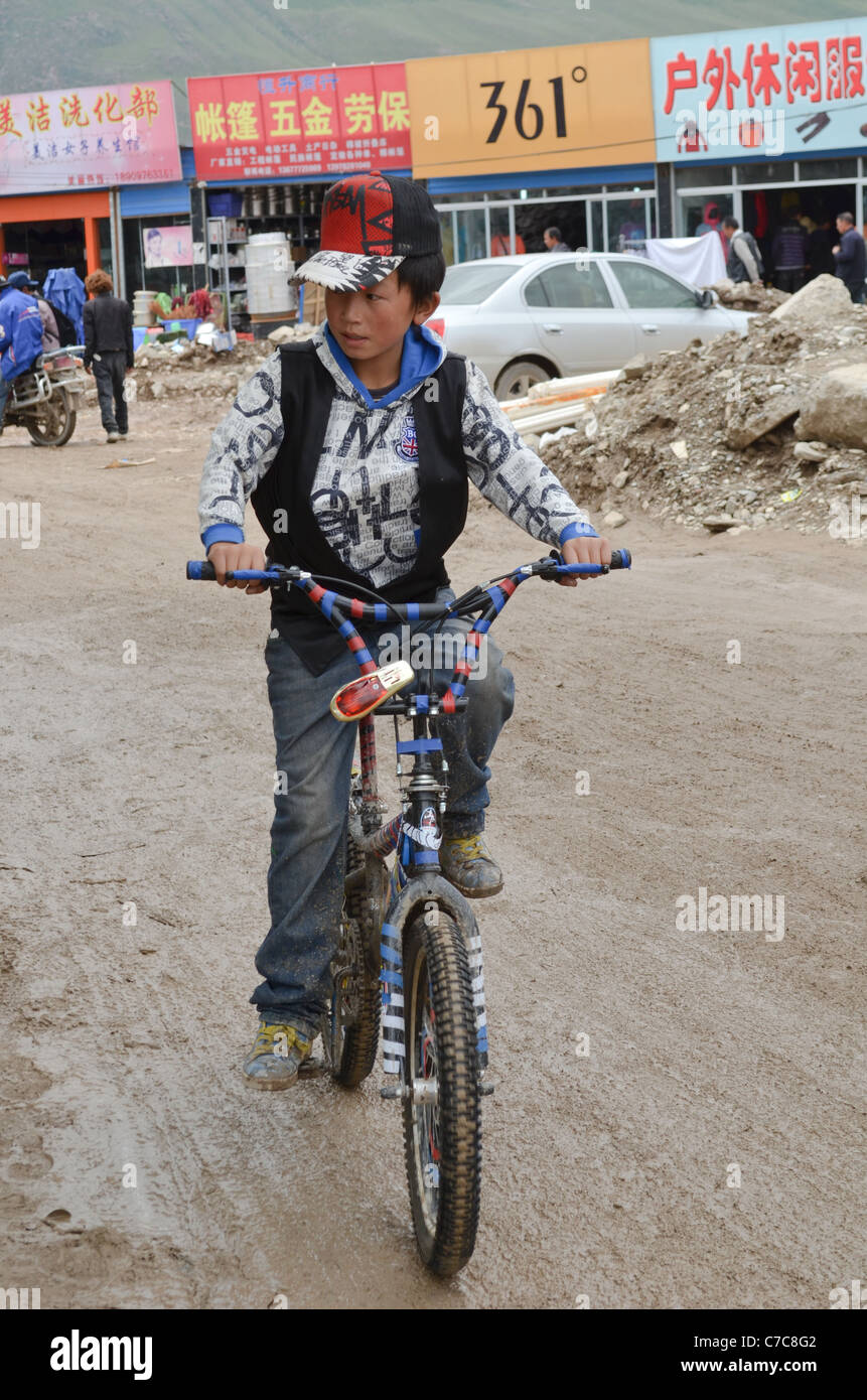 Un ragazzo di cicli su strade di fango tra negozi temporanei poiché un terremoto (2010) girato la maggior parte degli edifici in macerie, Yushu, Cina Foto Stock