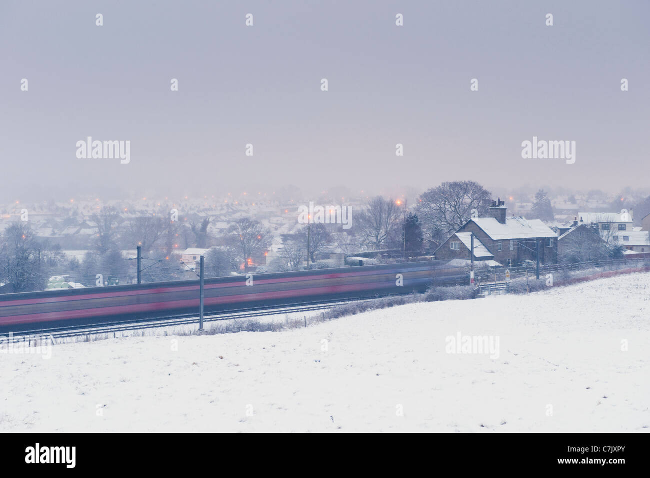Serata invernale innevata, treno passeggeri in velocità oltre villaggio rurale e campo coperto di neve bianca - Wharfedale Line, West Yorkshire, Inghilterra, Regno Unito. Foto Stock
