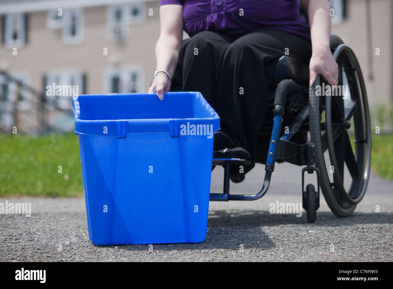 Donna con la spina bifida in una sedia a rotelle di prelevare il suo contenitore di riciclaggio presso la strada Foto Stock