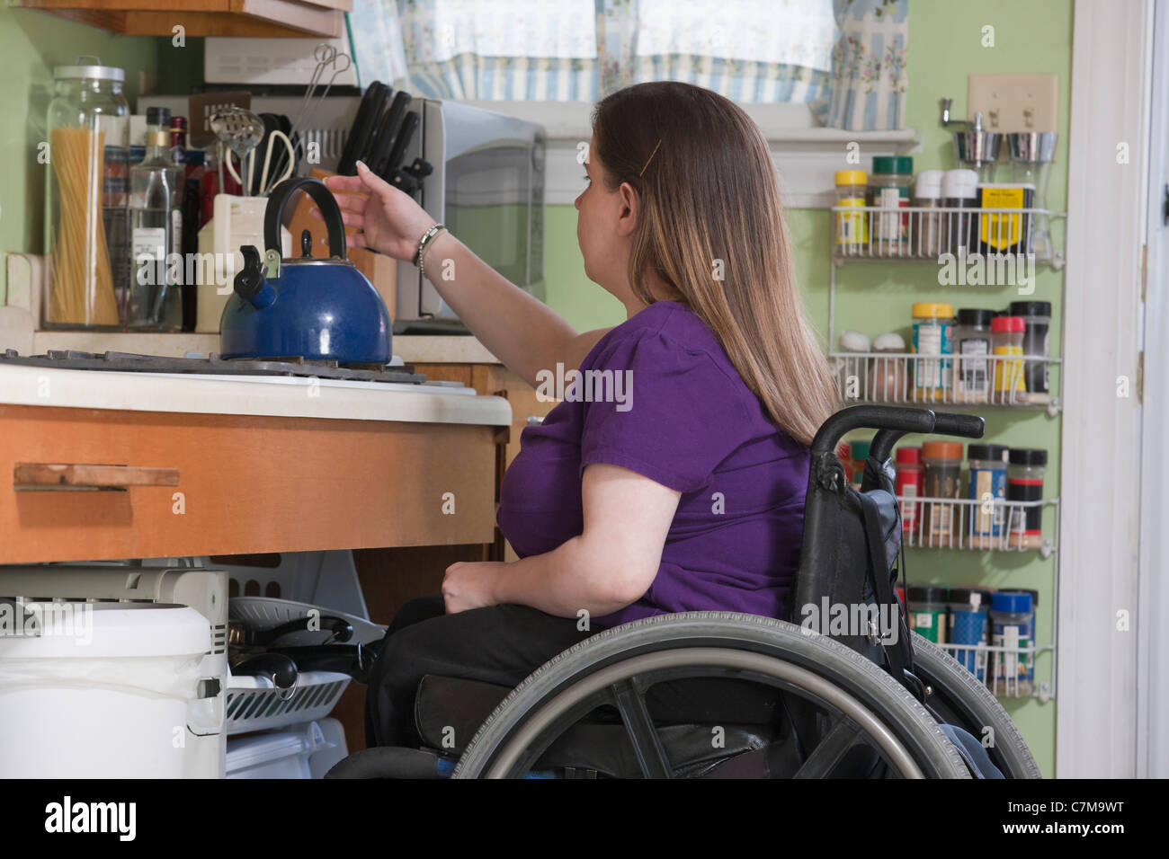 Donna con la spina bifida in una sedia a rotelle il posizionamento di un bollitore nella sua cucina accessibile Foto Stock