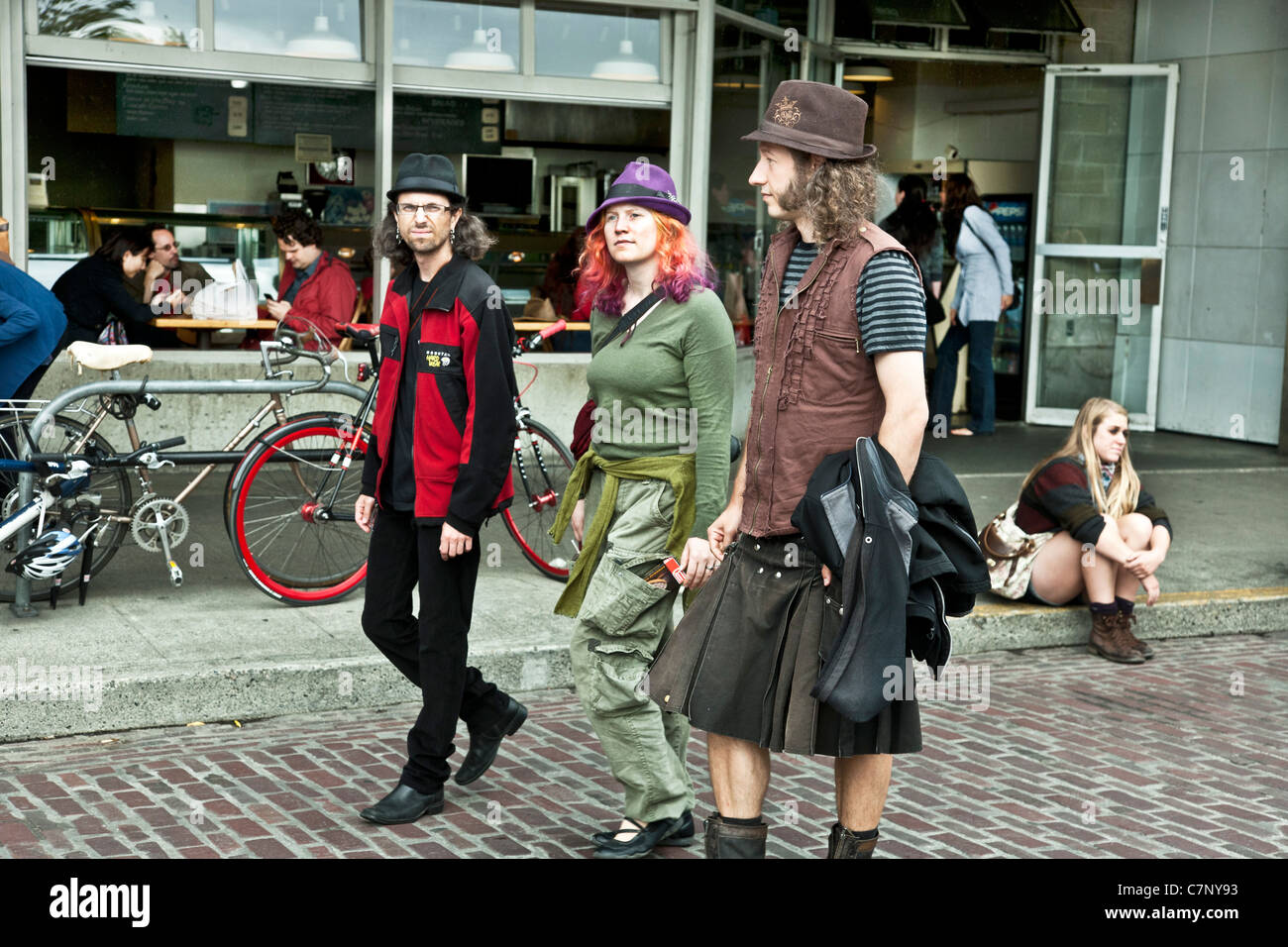 Ragazza con i capelli arancione e viola hat camminando su Pike Place accompagnato da 2 capelli lunghi giovani uomini uno indossando un kilt, in fieri display grunge di Seattle Foto Stock