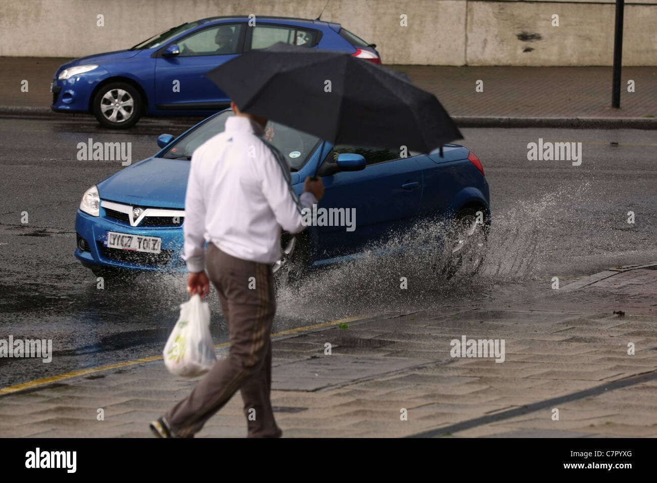 Un auto schizzi attraverso una pozzanghera allo stesso tempo come un uomo che porta un ombrello passa Foto Stock