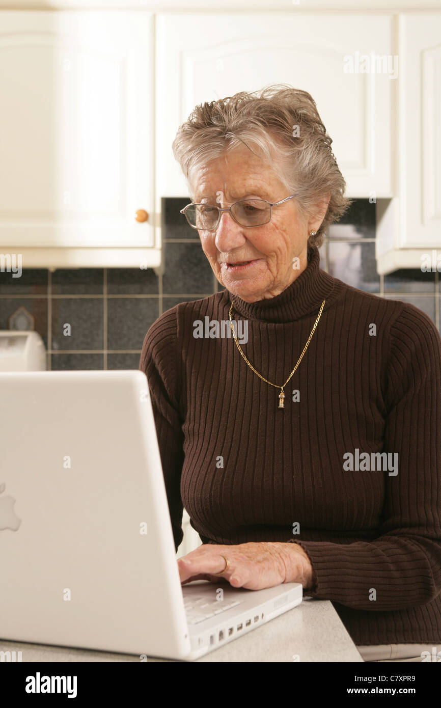 Una donna nella sua metà anni ottanta usando un computer portatile nella sua cucina. Foto Stock