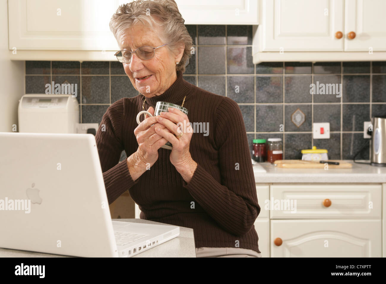 Una donna nella sua metà anni ottanta usando un computer portatile nella sua cucina. Foto Stock