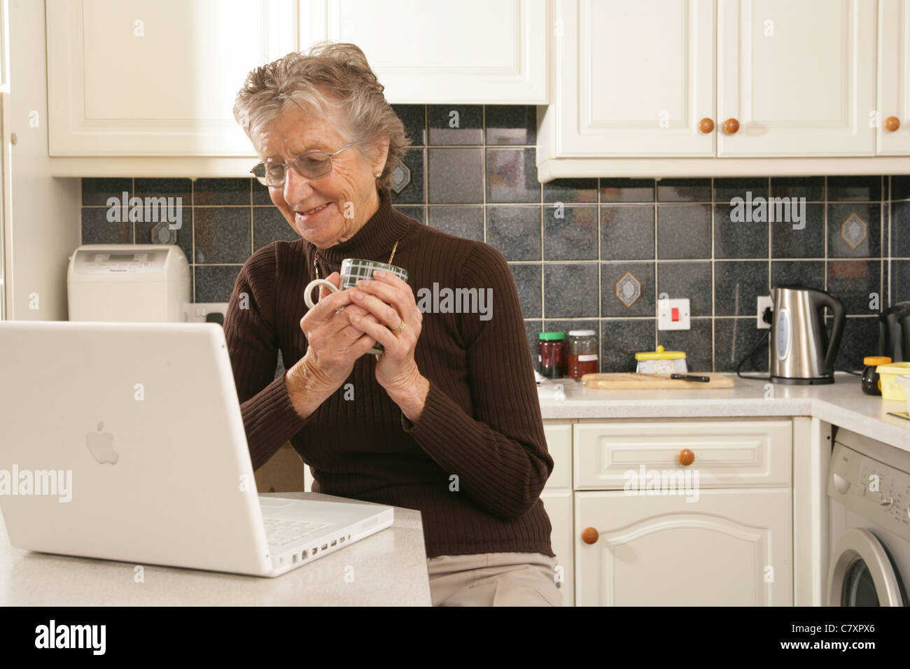 Una donna nella sua metà anni ottanta usando un computer portatile nella sua cucina. Foto Stock
