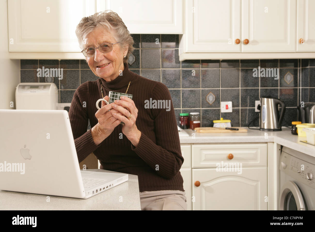 Una donna nella sua metà anni ottanta usando un computer portatile nella sua cucina. Foto Stock