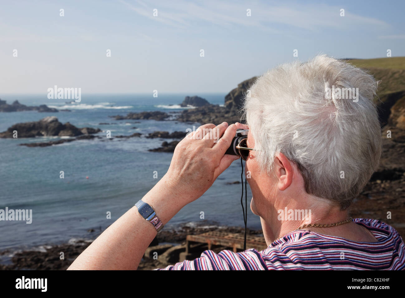 Senior donna pensionato guardando attraverso il binocolo al mare sulla costa di più a sud il punto sul territorio continentale del Regno Unito. Lizard Point Cornwall Inghilterra REGNO UNITO Foto Stock