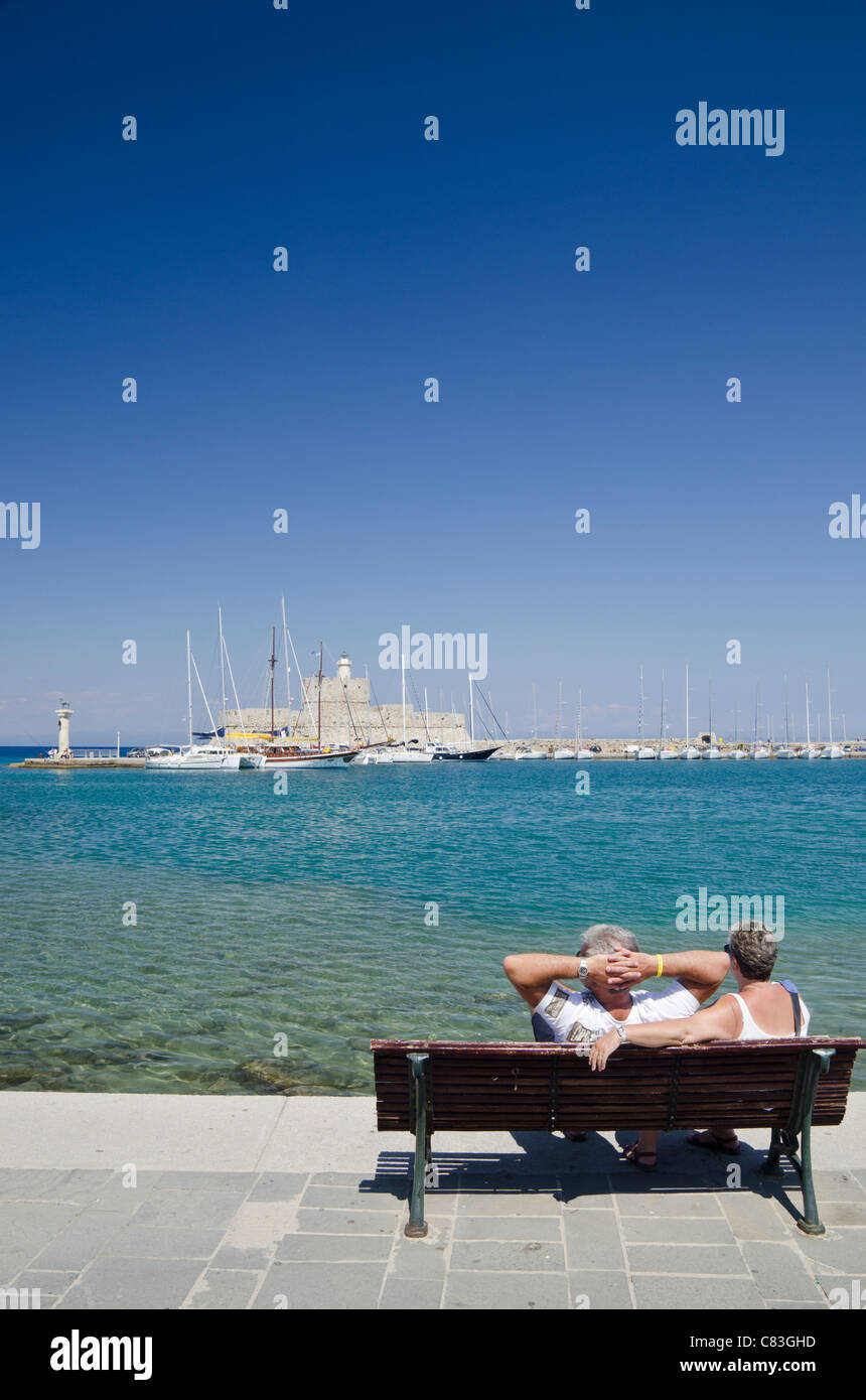 Un paio di rilassarvi su una panchina sul lungomare che guarda verso la Fortezza di San Nicola in Mandraki Harbour, Rodi, Rodi, Grecia Foto Stock