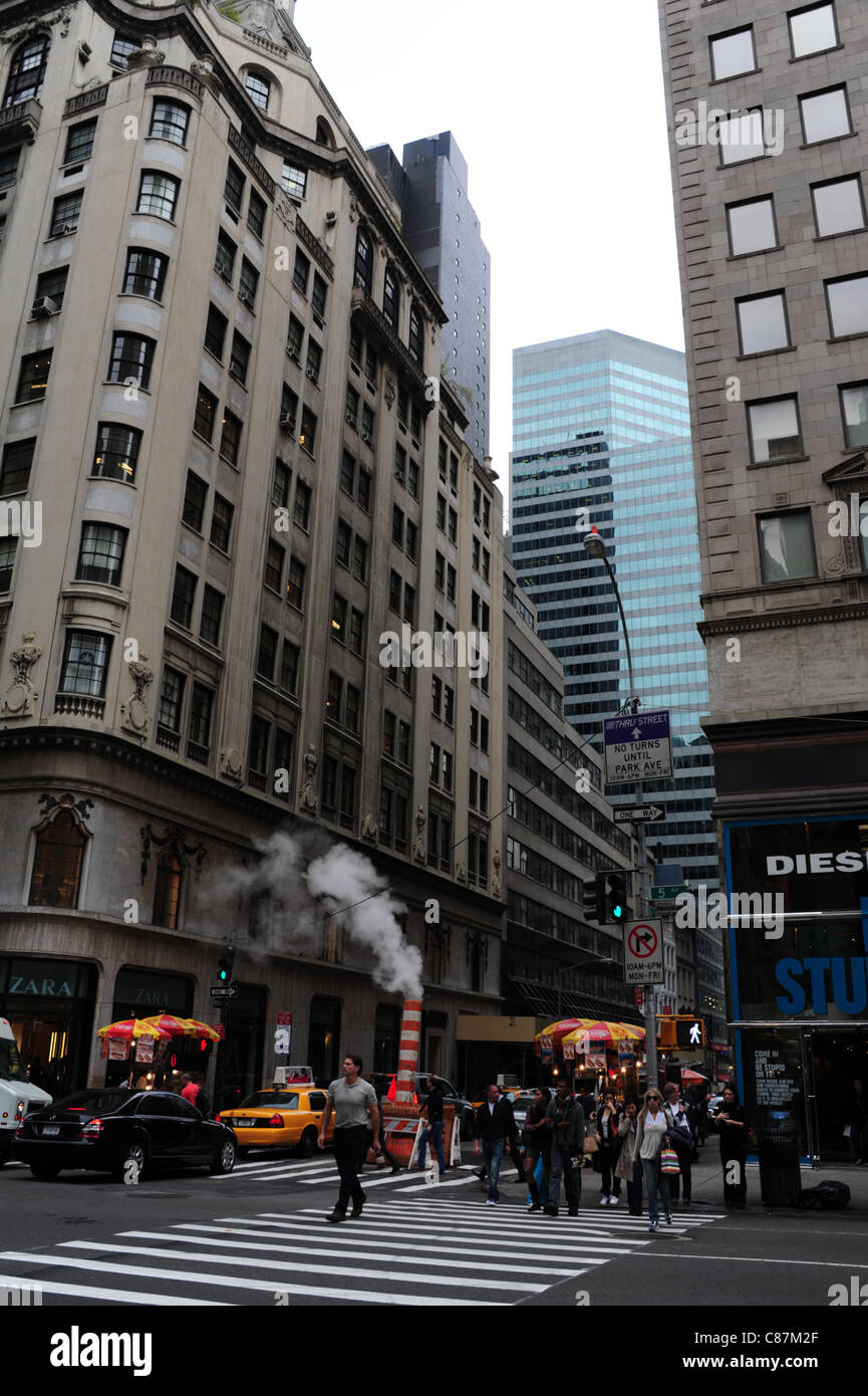 Le persone che attraversano la Quinta Avenue, con il riscaldamento del vapore acqueo passando da un rosso camino in plastica presso East 54th Street Junction, New York City Foto Stock