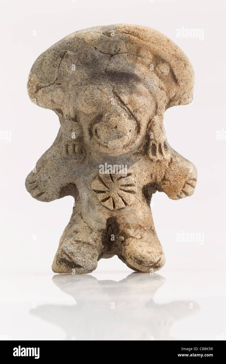Una piccola, antica statuetta in terracotta si erge in posizione verticale su una superficie bianca riflettente. La figura ha una testa arrotondata, simile a funghi, con linee incise che suggeriscono caratteristiche facciali, e un'ampia, semplificata Foto Stock