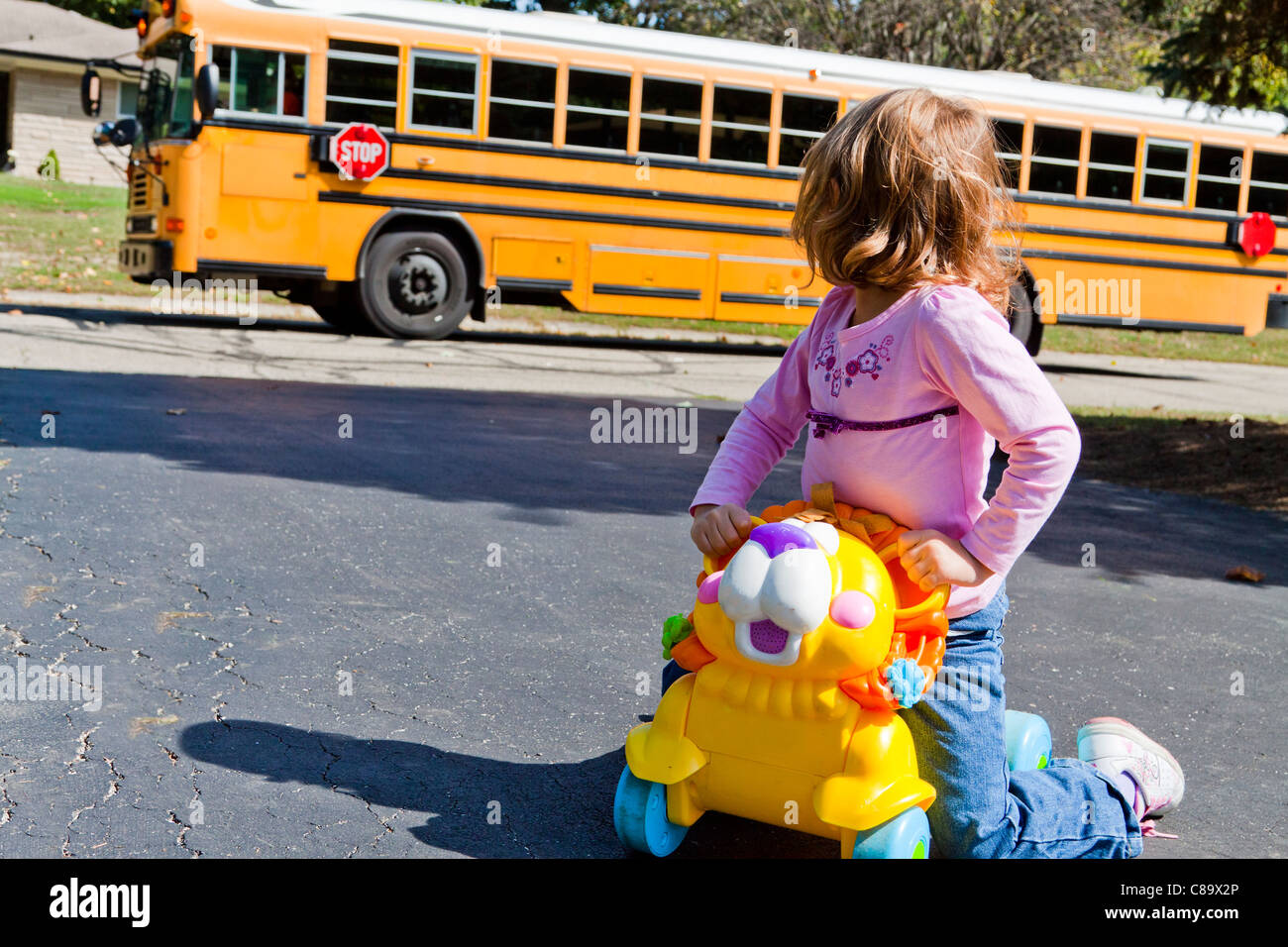 A quattro anni e mezzo che bambina si siede su un cavallo giocattolo in suo passo carraio si volta a guardare un giallo scuola bus che passa. Foto Stock