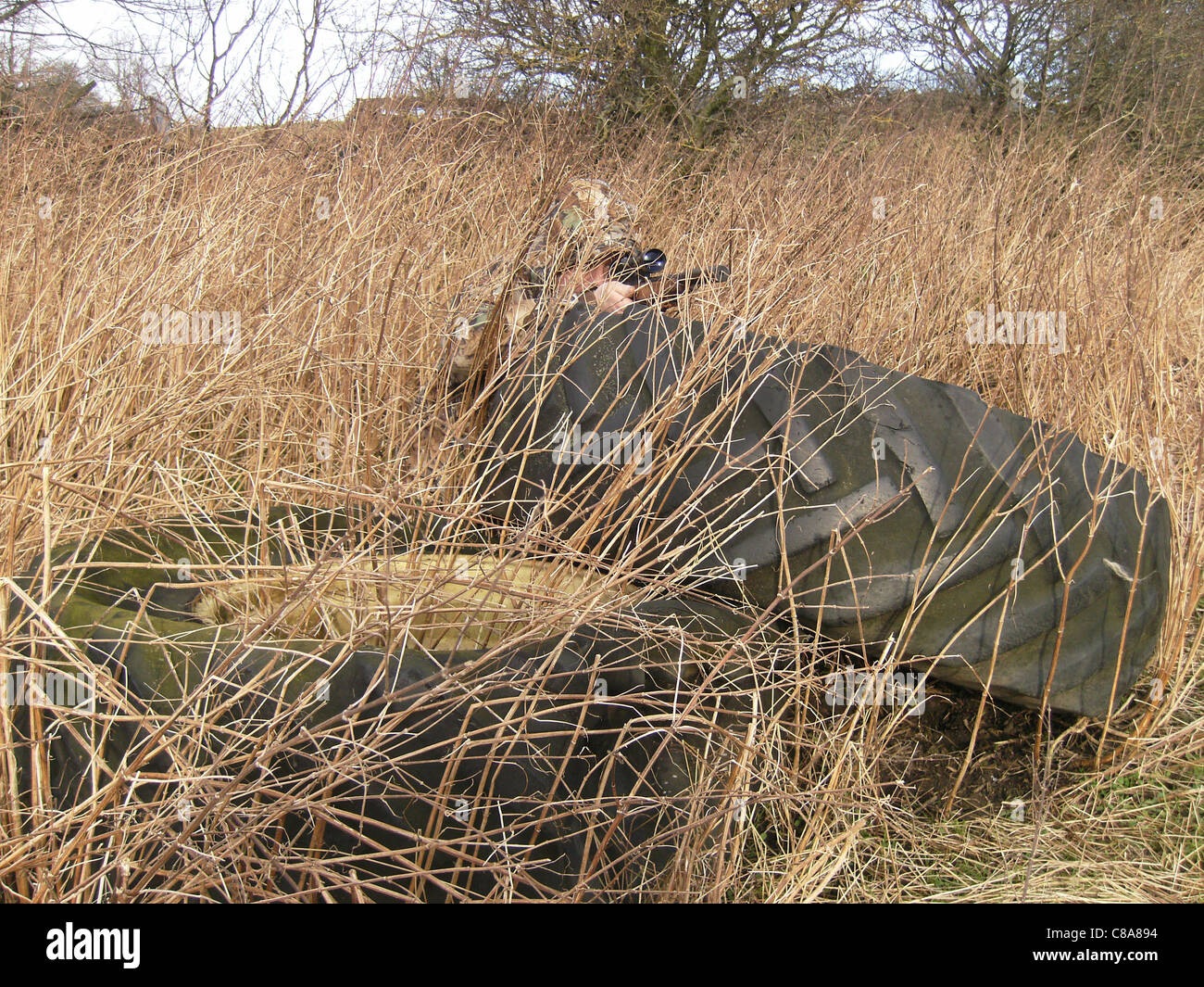 Un cacciatore accovacciato verso il basso dietro il vecchio trattore pneumatici in Abbigliamento camouflage in erba lunga tenendo un fucile e guardando attraverso la portata in weardale. Foto Stock