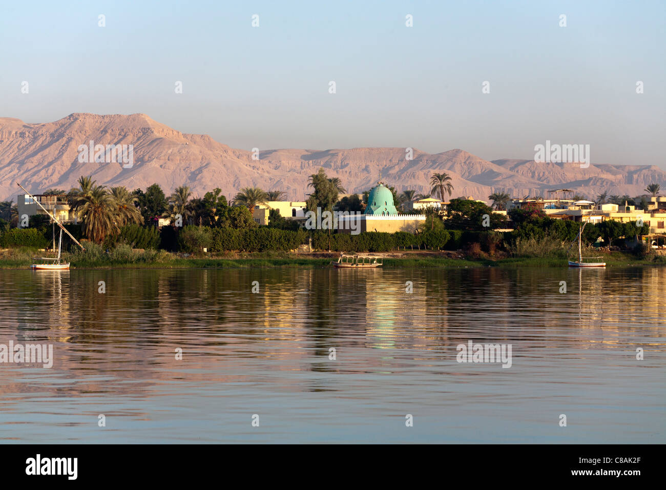 La sezione del fiume Nilo banca con alberi e moschea e la West Bank montagne sullo sfondo illuminato da golden luce della sera Foto Stock
