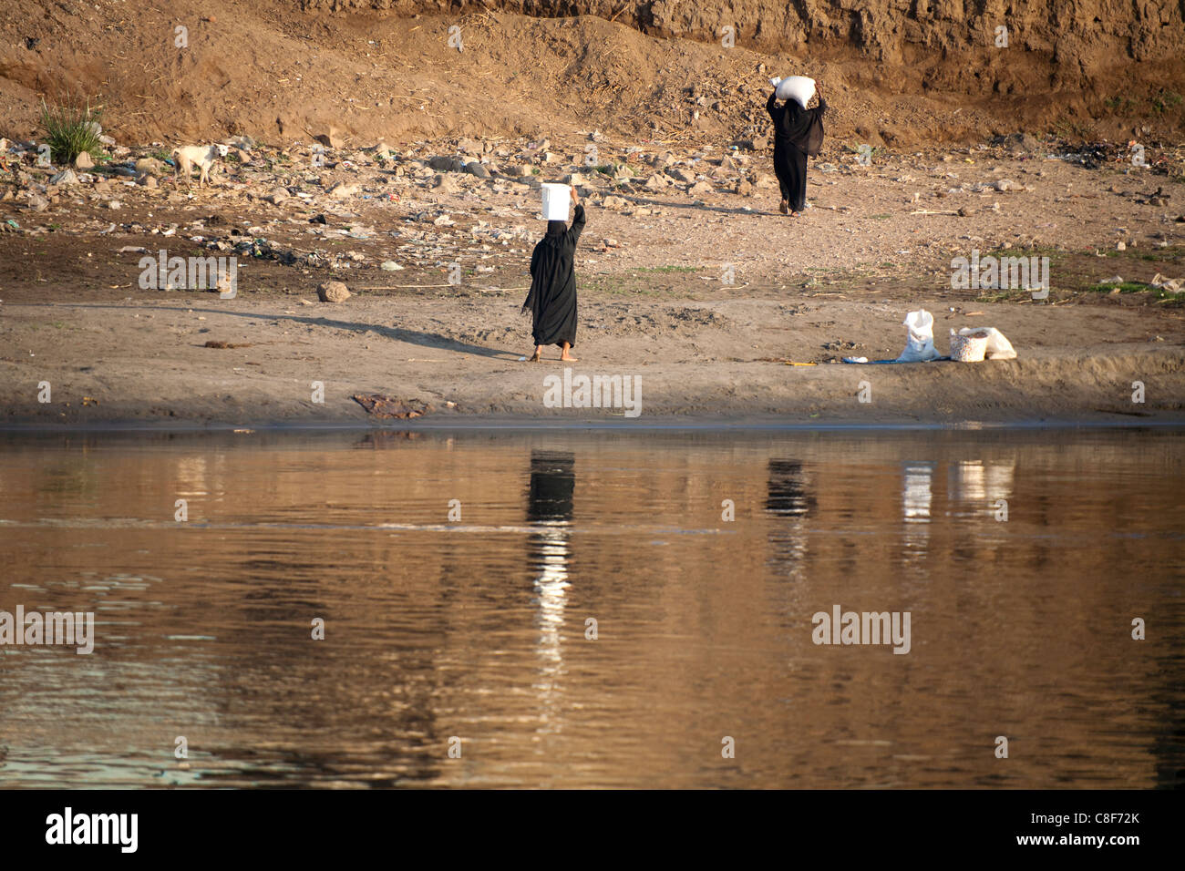 Una sezione del fiume Nilo e banca di fango con due donne in nero che trasportano contenitori bianchi sulle loro teste riflessa nell'acqua ancora Foto Stock