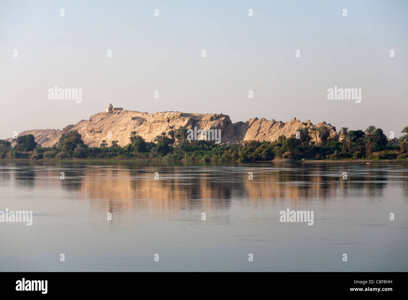 La sezione del fiume Nilo banca con affioramento roccioso con il santuario in cima e alberata base tutti riflette i morti in acqua calma Foto Stock