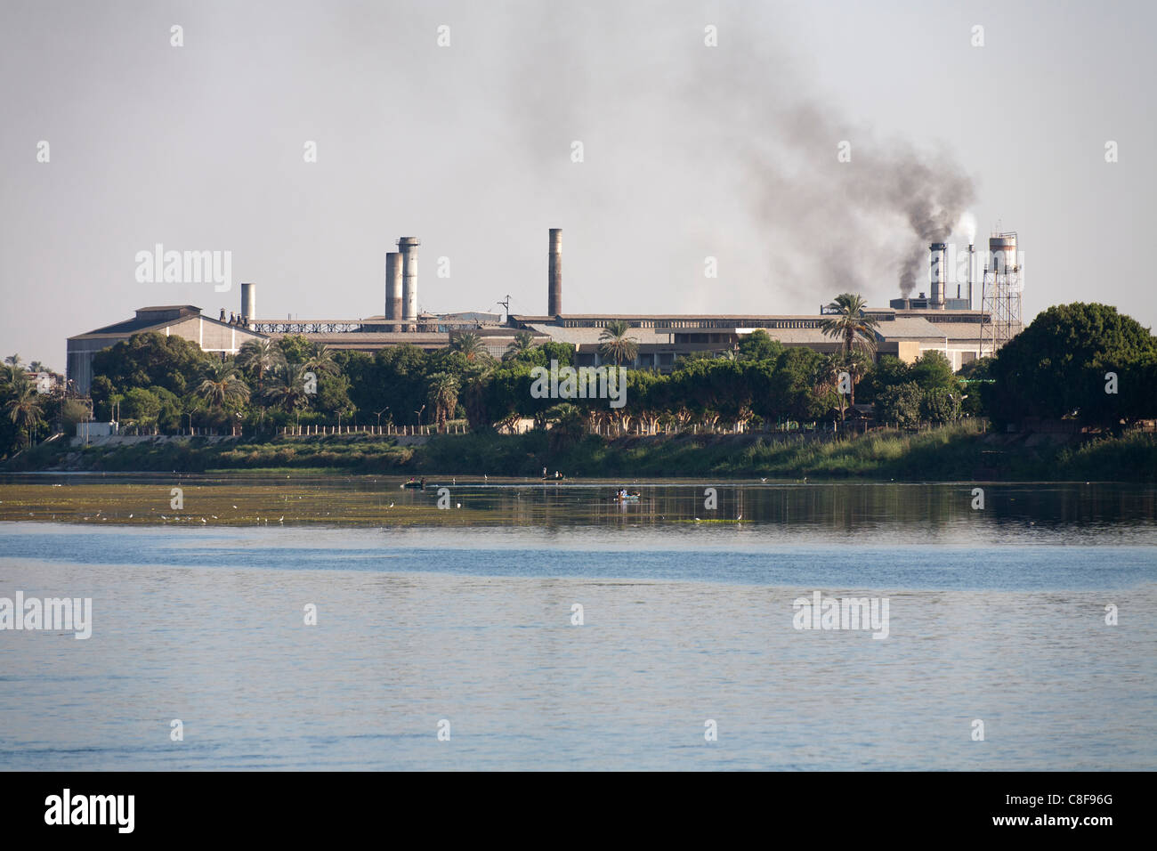 Una sezione del fiume Nilo banca con impianto industriale con comignoli fumanti dietro alberata in banca e numerose piccole barche da pesca Foto Stock