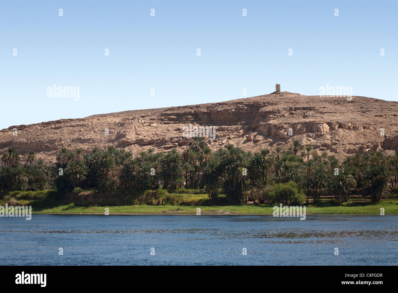 I resti di una torre in pietra su un lontano deserto roccioso tumulo sulle rive del Nilo, una linea di alberi in primo piano e il fiume nella parte anteriore Foto Stock