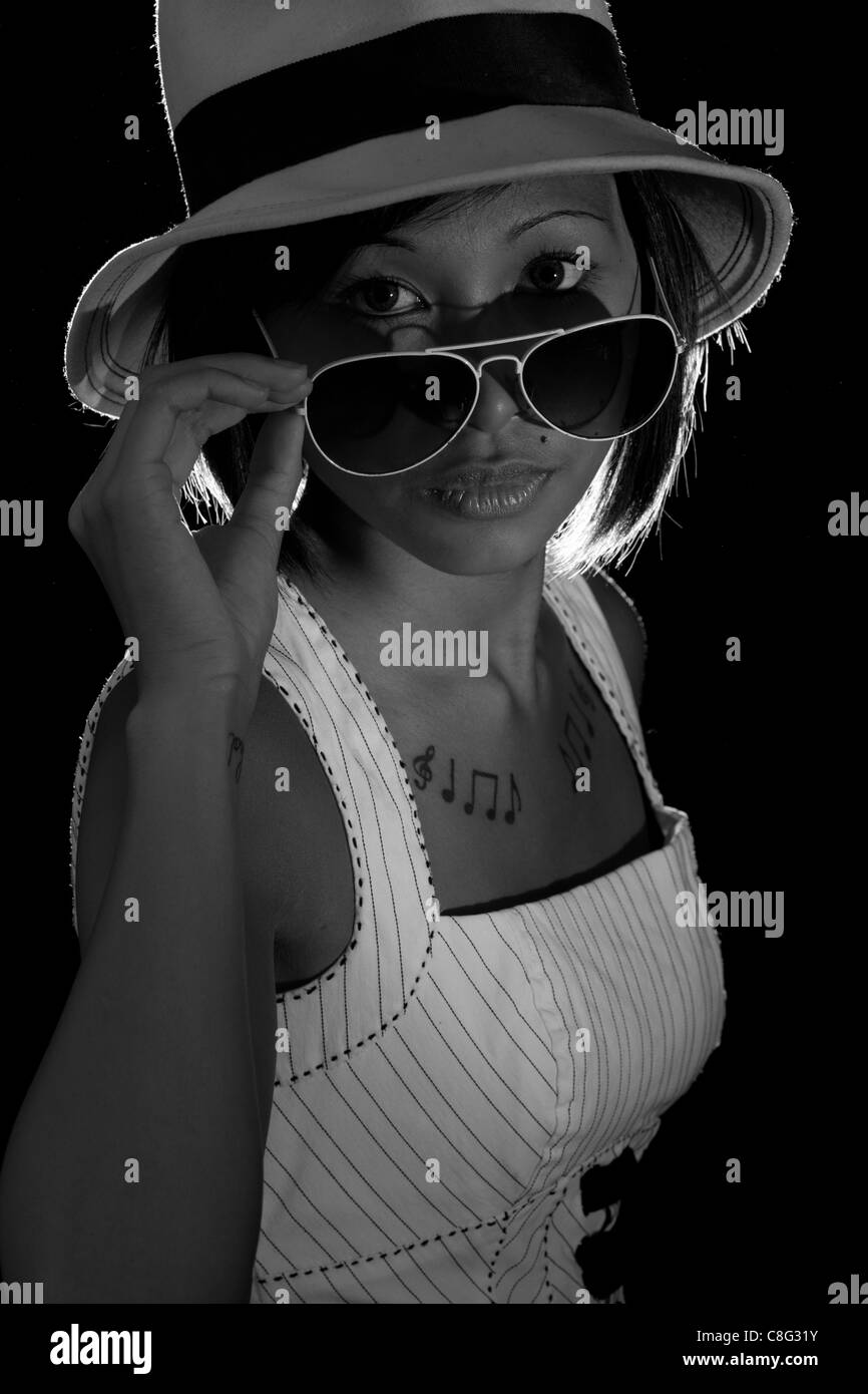 Una immagine illuminata da dietro di una bella ragazza asiatica indossando occhiali da sole e cappello. L'immagine è in bianco e nero. Foto Stock