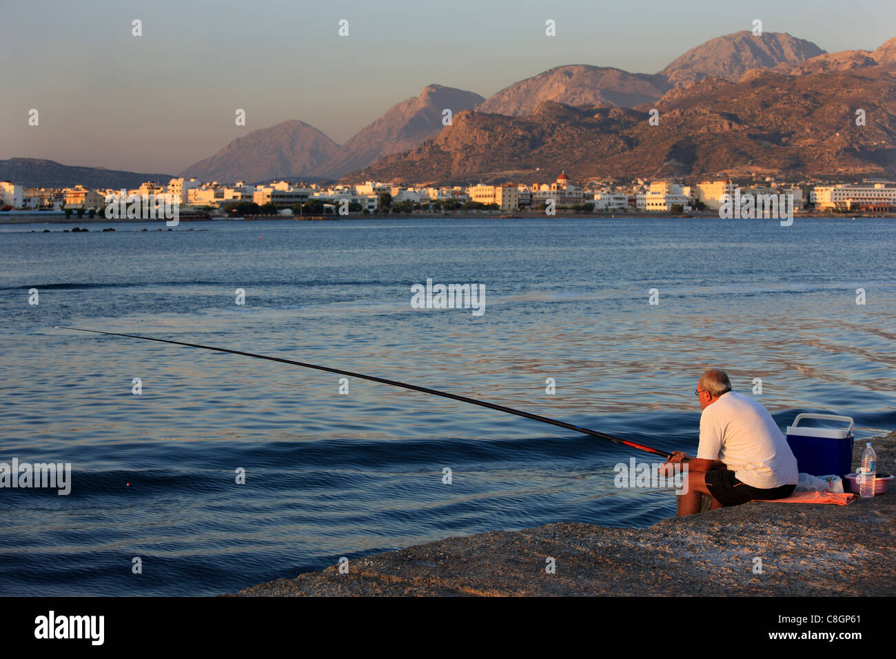 Un uomo solitario pesca, seduto sulla scogliera, di fronte alla città di Ierapetra, Lassithi, Creta, Grecia Foto Stock
