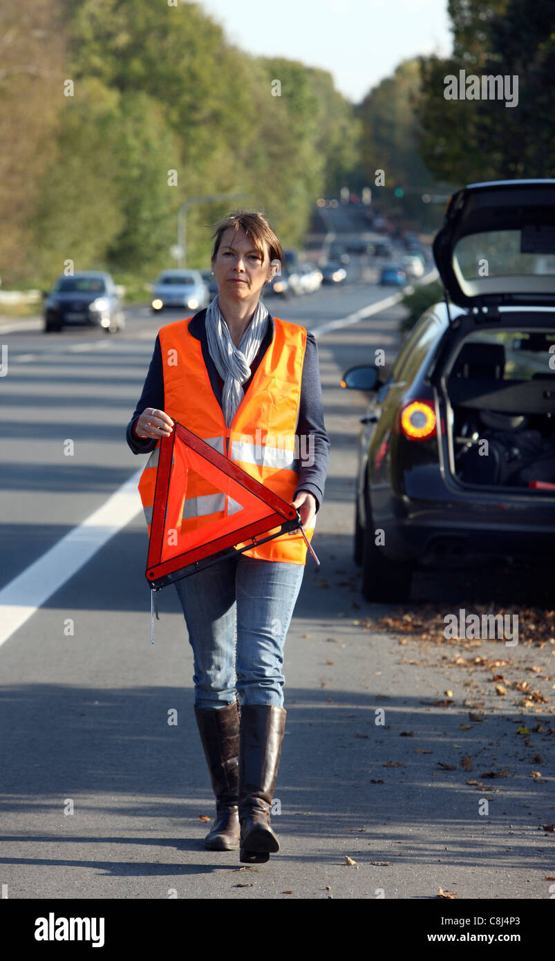 Ripartizione per auto, su una autostrada, driver femminile, indossa una divisa da lavoro ad alta visibilità, impostare un triangolo di avvertenza. Foto Stock