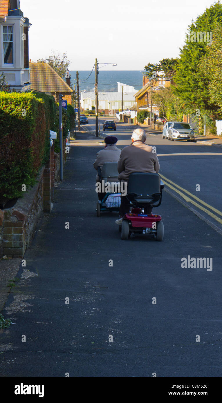 Due anziani ride scooter di mobilità lungo il marciapiede verso il mare. Foto Stock