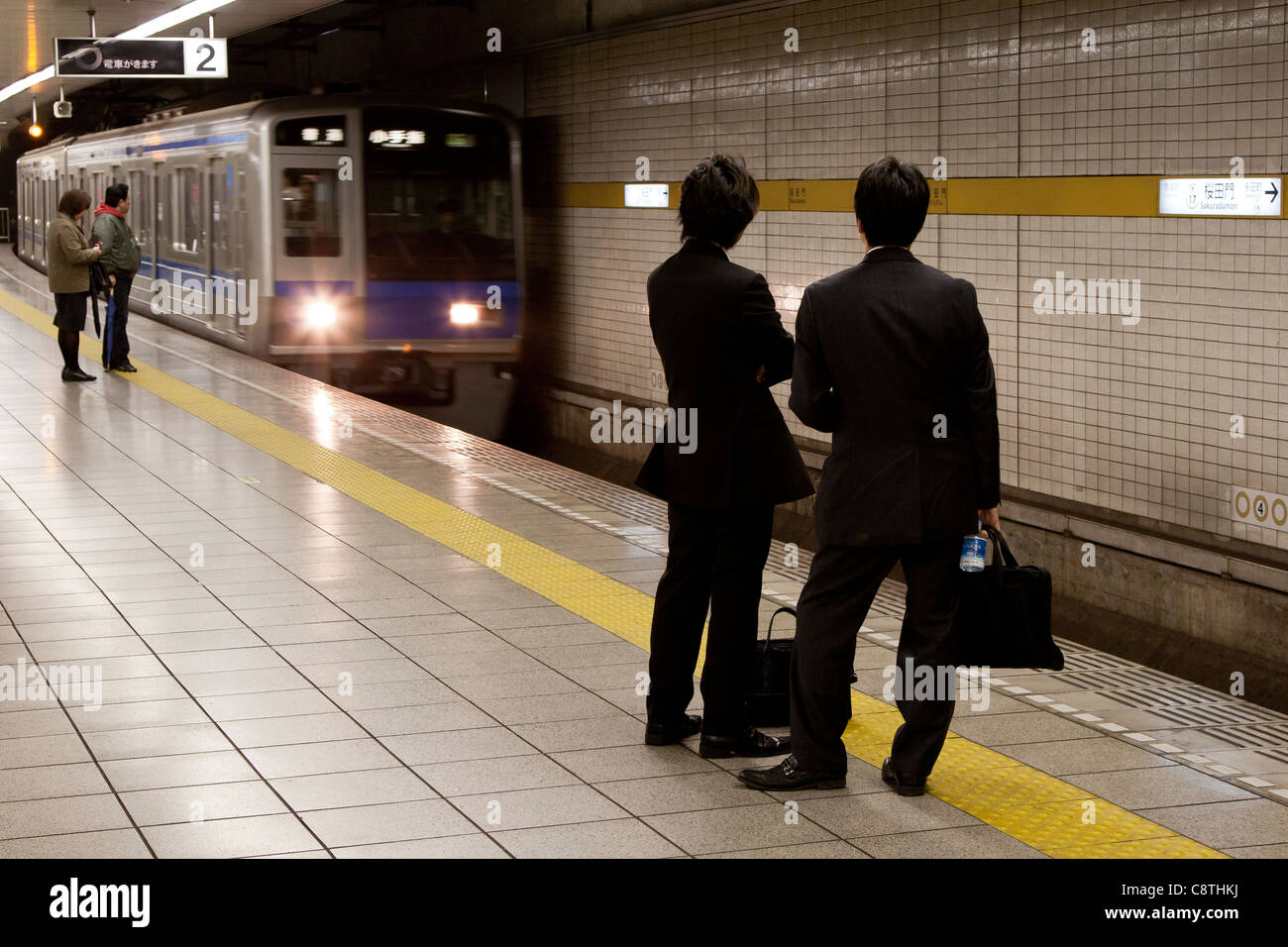 Uomini d'affari giapponesi o salarymen attendere su una piattaforma a stazione di Sakuradamon come un " commuter " treno arriva. Tokyo, Giappone. Foto Stock