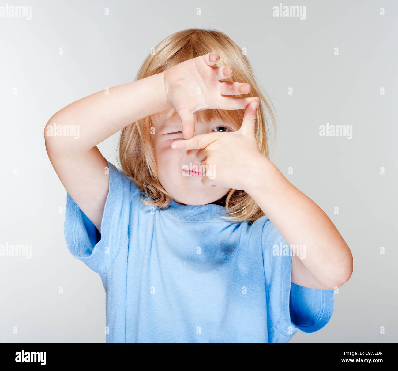 Little Boy con lunghi capelli biondi guardando attraverso un telaio di dito - isolato su grigio Foto Stock