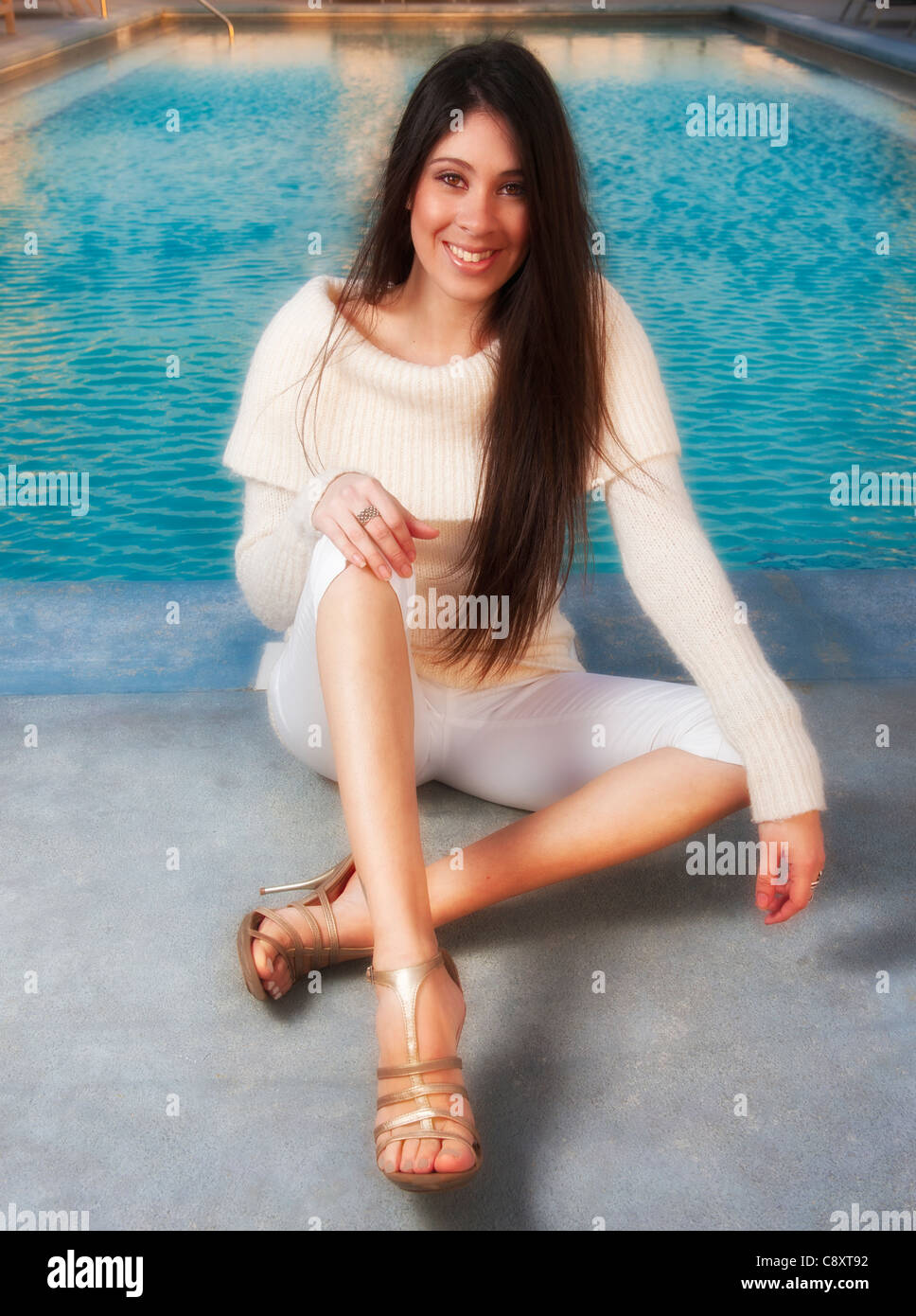 Attraente giovane donna ispanica 20s si siede a bordo piscina nel  pomeriggio indossando un lussuoso bianco / maglione beige, pantaloni capri  tacchi Foto stock - Alamy