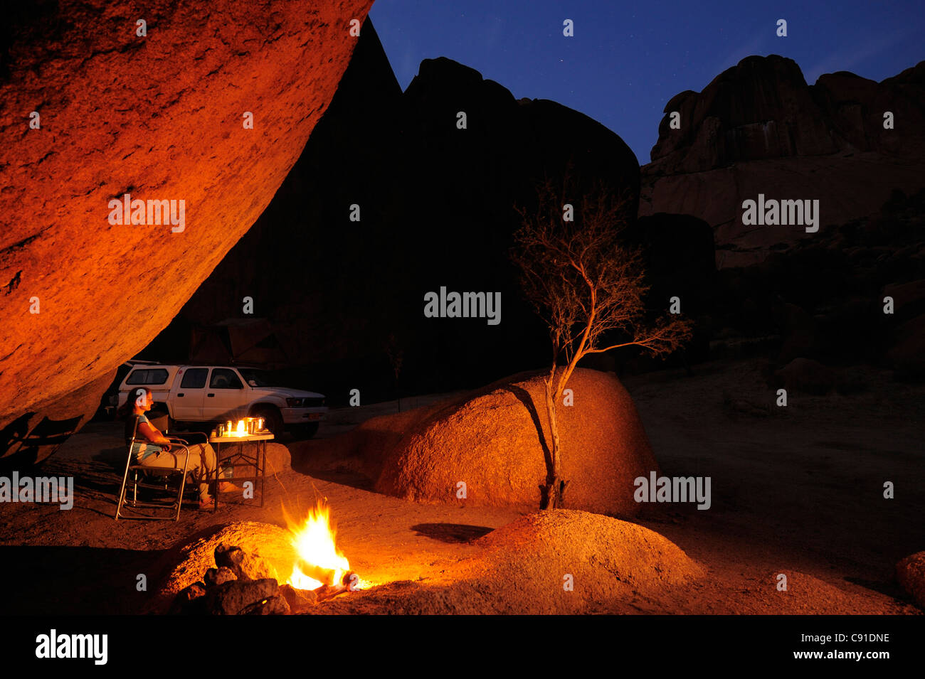 Donna seduta di fronte a barbecue il fuoco sotto la sporgenza di roccia, auto in background, grande Spitzkoppe, Namibia Foto Stock