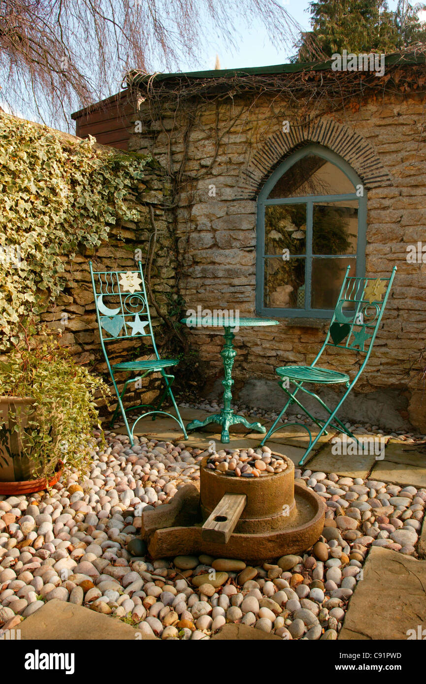 Sedie in metallo e tavolo con ciottoli e pietre area patio con fontana e giardino esterno casa sul giardino inglese. Foto Stock