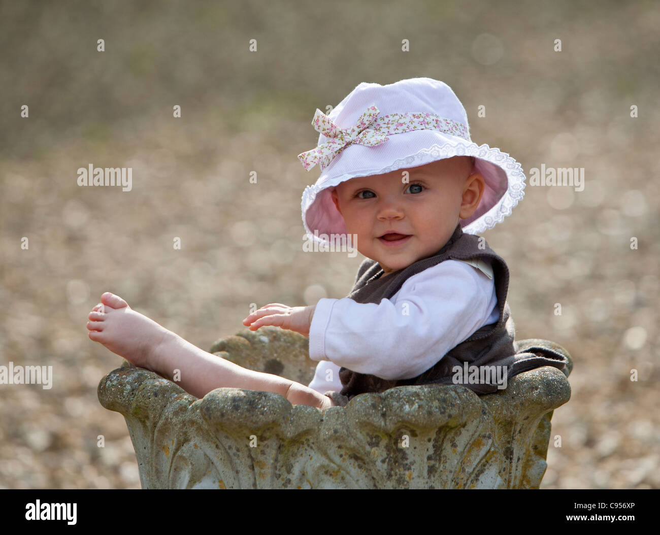 Baby ragazza seduta in una piantatrice di pietra che indossa un cotone cappello per il sole e sorridere per la fotocamera Foto Stock