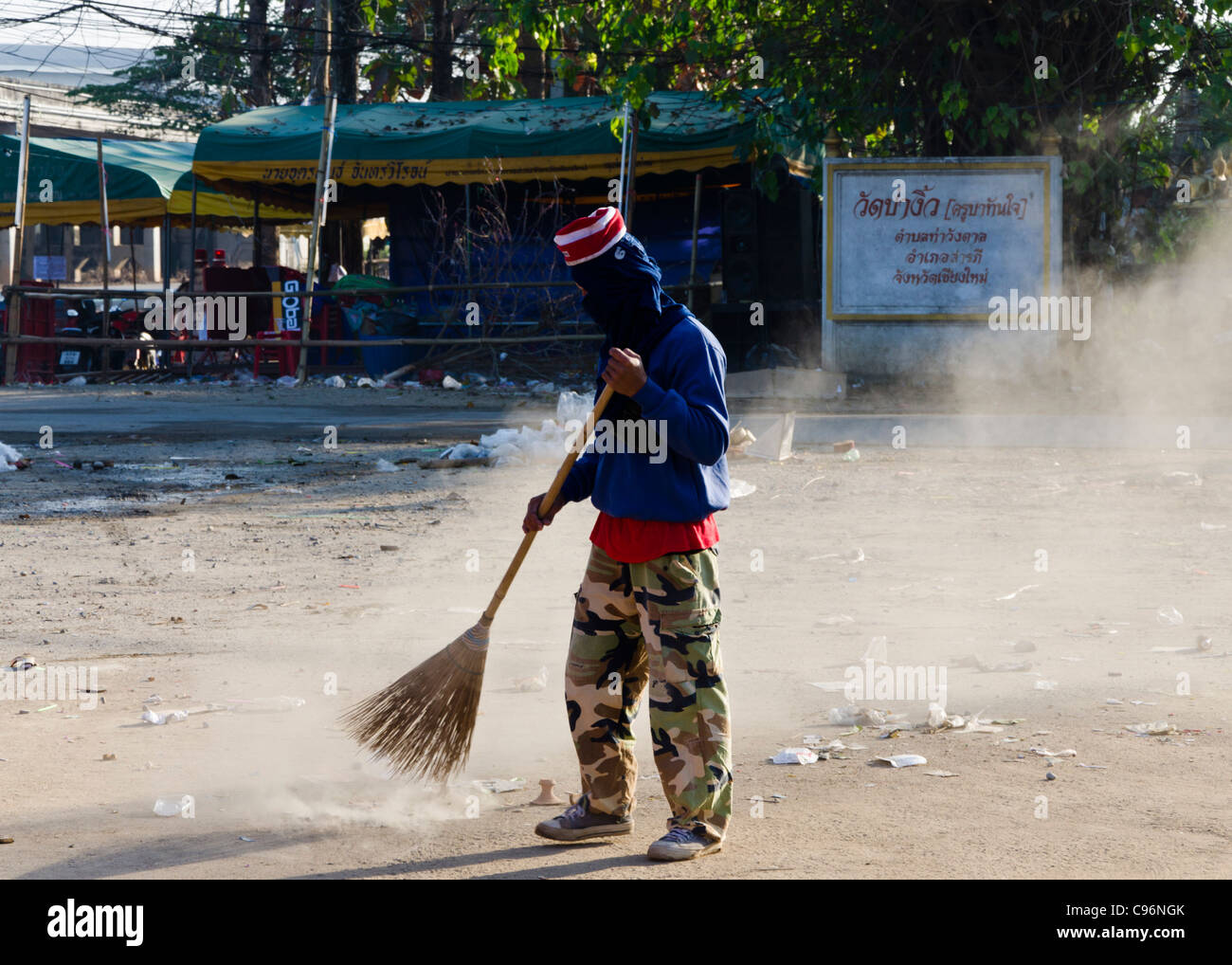 Giovane uomo con maschera facciale e Pantalone mimetico spazzando via i rifiuti con grande scopa di paglia dopo festival nel nord della Thailandia Foto Stock