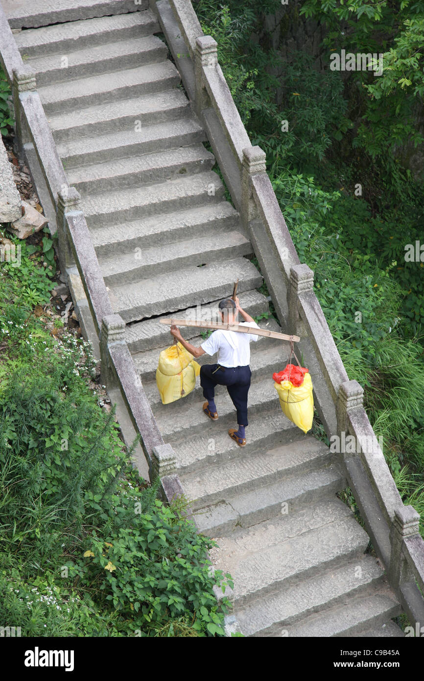 Un uomo con un palo portante caricate con sacchetti utilizza un bastone da passeggio mentre salendo le scale presso il monte Jiuhua, Cina. Foto Stock