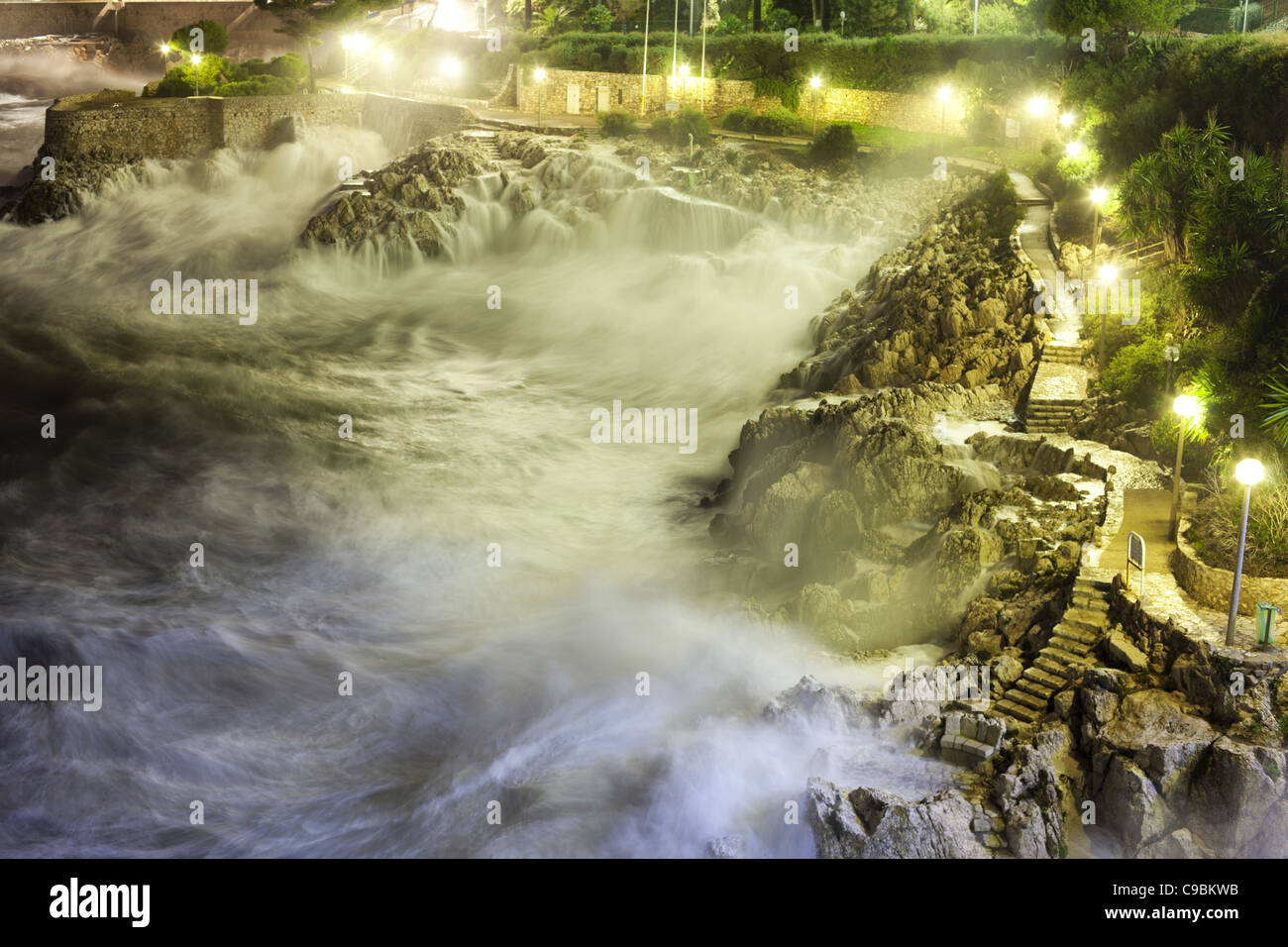 Esposizione notturna di un mare accidentato che si schiantano su una costa rocciosa lungo un sentiero illuminato. Nizza, Costa Azzurra, Francia. Foto Stock